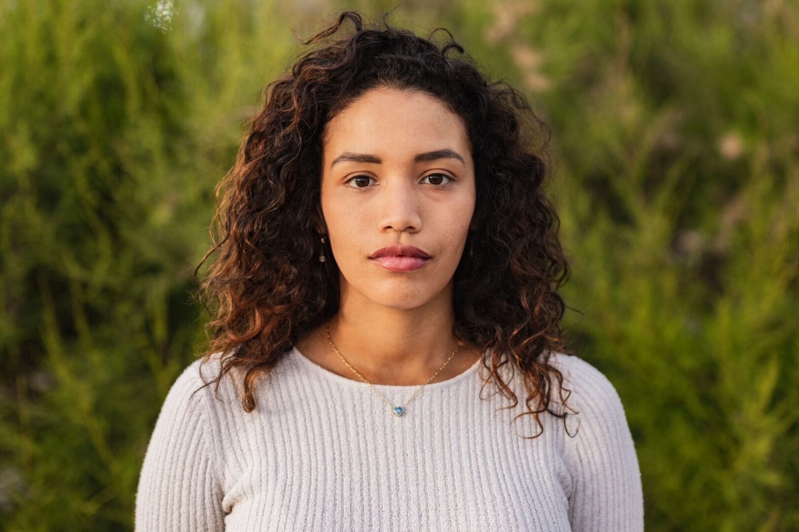 A person with curly hair and a neutral expression stands in front of greenery, wearing a light-colored sweater and a necklace.