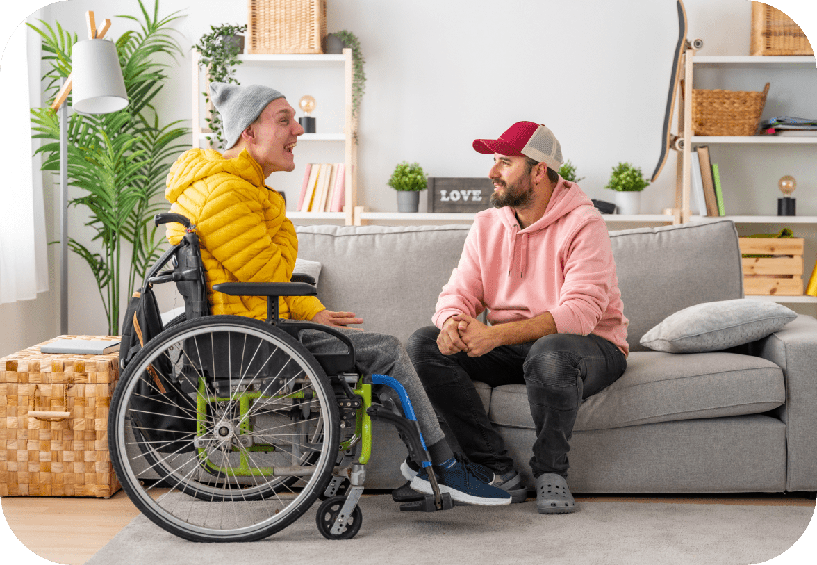 Two men are engaged in conversation in a living room. One is seated in a wheelchair wearing a yellow jacket, and the other sits on a sofa wearing a pink hoodie and red cap.