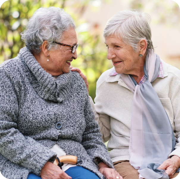 Two elderly women sit together outdoors, smiling and talking, with one resting a hand on the other's shoulder.
