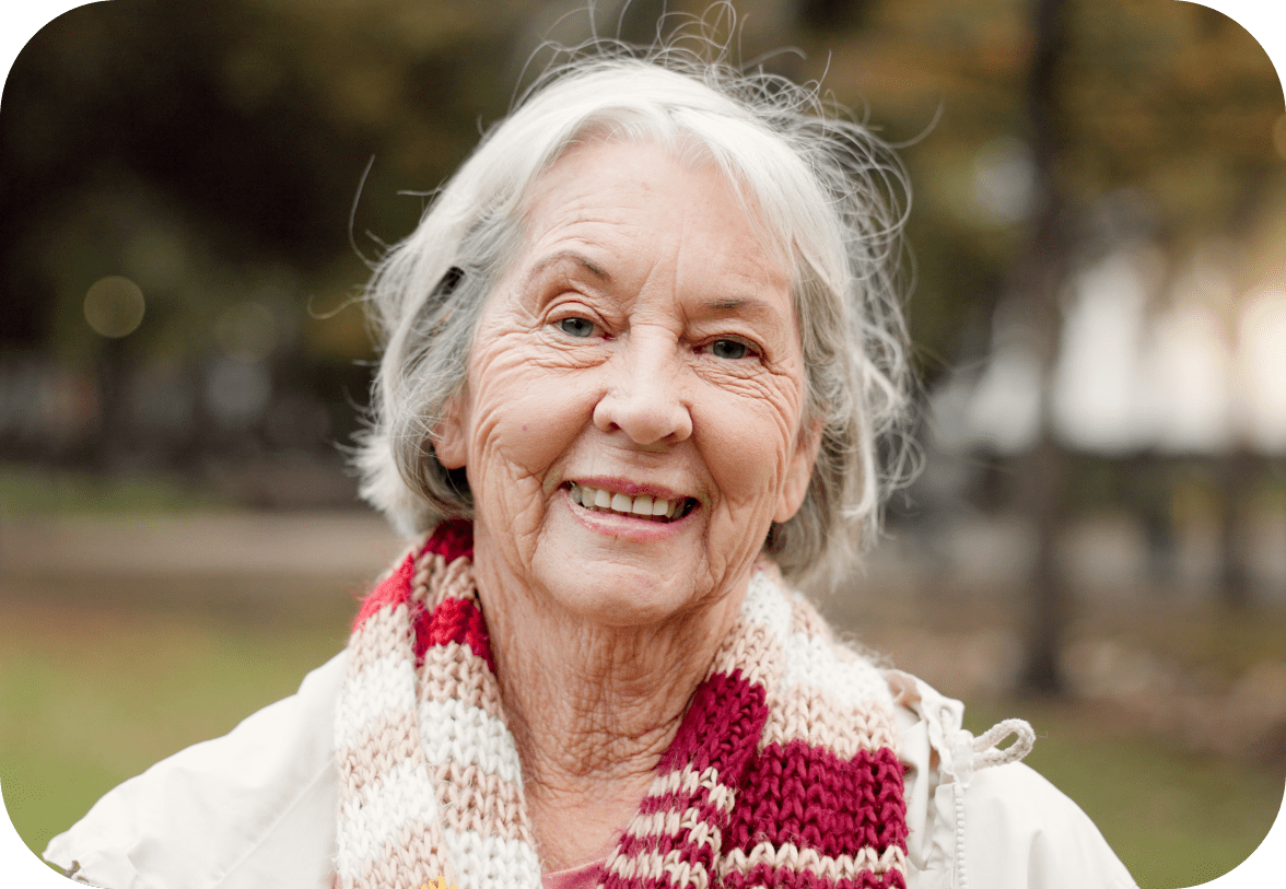 An elderly woman with gray hair smiles while wearing a colorful scarf. She stands outdoors with a blurred background.