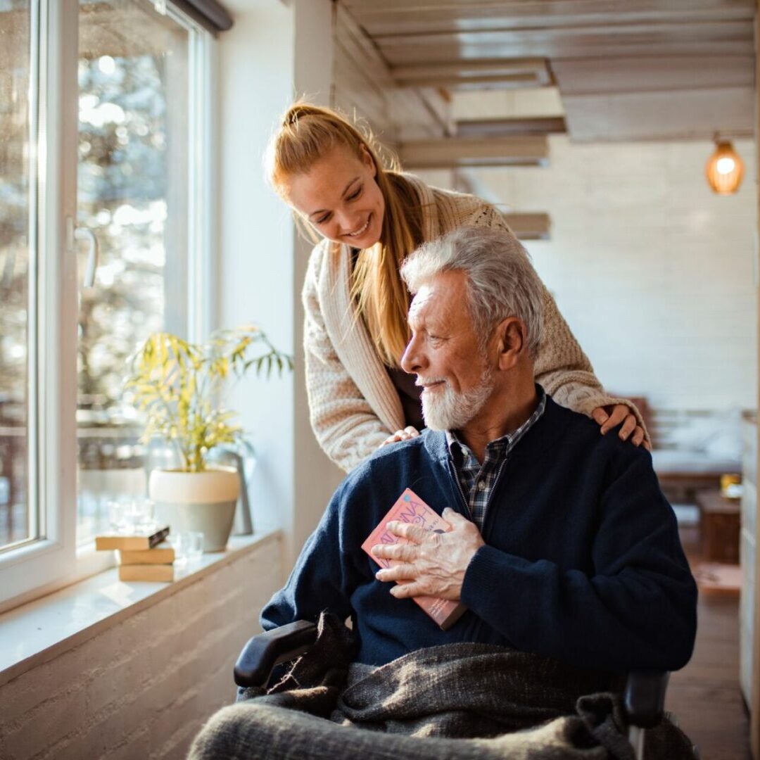 An elderly man in a wheelchair holds a card, smiling up at a woman standing beside him near a sunny window.