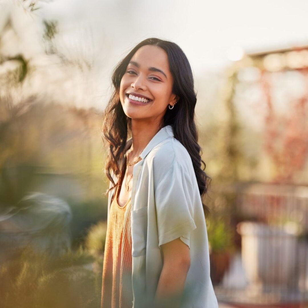A woman with long black hair smiles while standing outdoors in a garden setting. She wears a light jacket over a brown top.