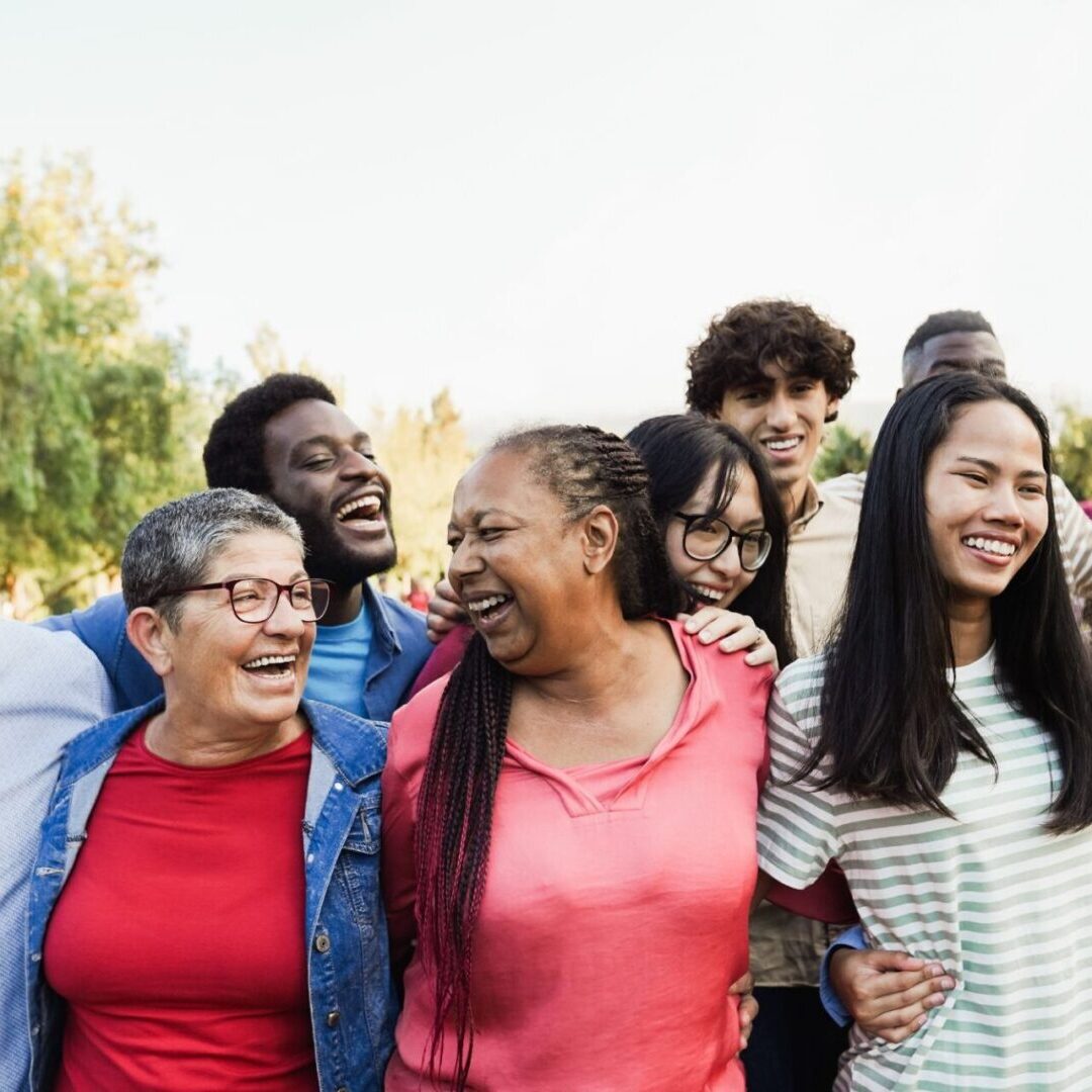 A diverse group of people stands together outdoors, smiling and laughing, with trees in the background.